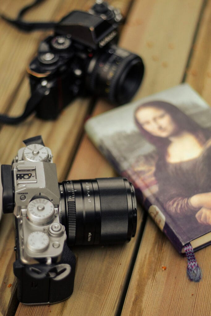 Two vintage cameras and a notebook with the Mona Lisa cover rest on a wooden table. The scene conveys a nostalgic, artistic vibe.