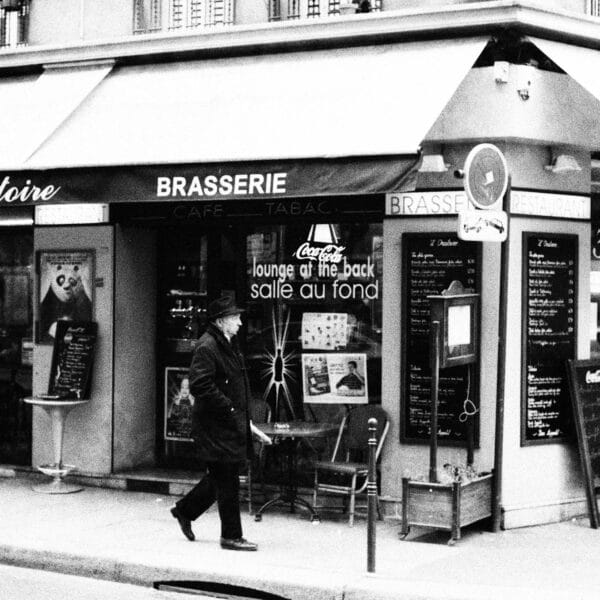 Black-and-white photo of a Parisian brasserie on a street corner. A man in a coat and hat walks by. The café has chairs outside and vintage signage, evoking a nostalgic, classic Paris atmosphere.