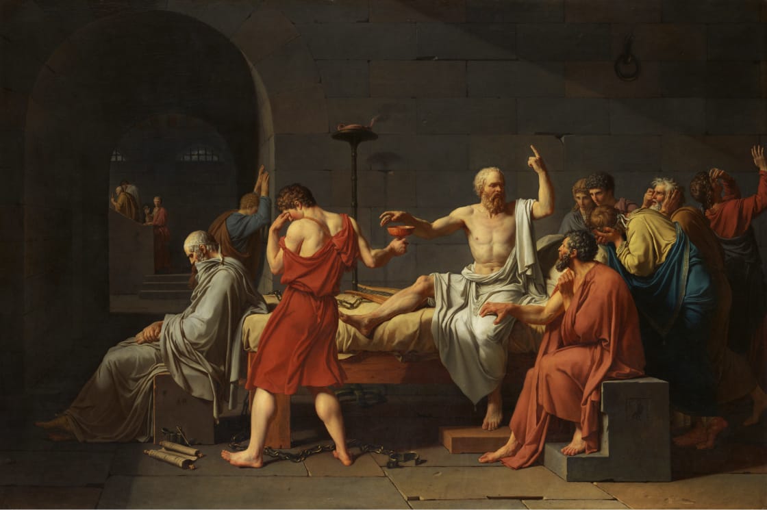 A group of men gather around an elderly, bearded man seated on a bed, reaching for a cup. The scene is set in a dim, stone-walled room, and the mood is somber as others react emotionally.