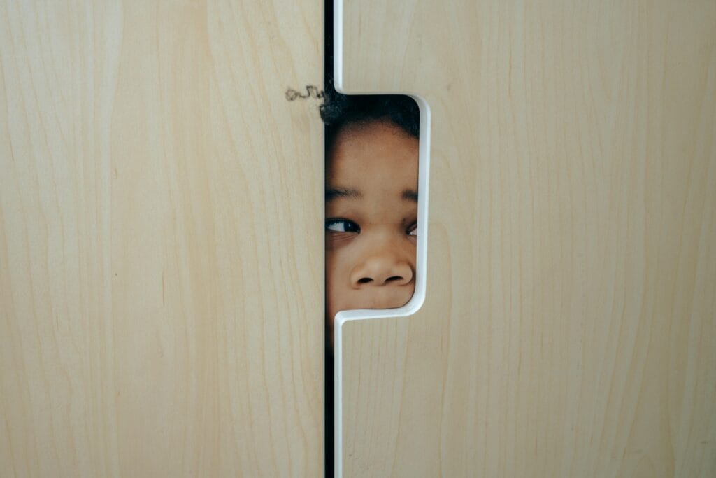 Hidden Object Puzzles. Young child peeking through a narrow opening in a light wooden door, showing only their eyes and part of their face.
