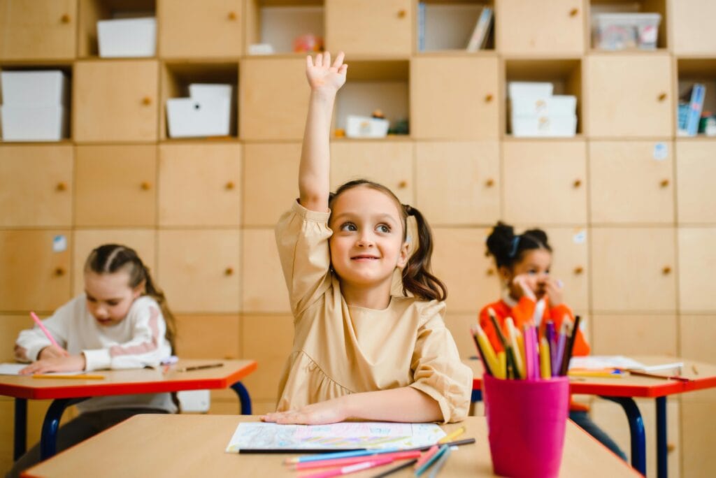 Spot the Difference. Young girl in a classroom raising her hand enthusiastically, with colorful pencils and drawings on the desk, while two other children work in the background.