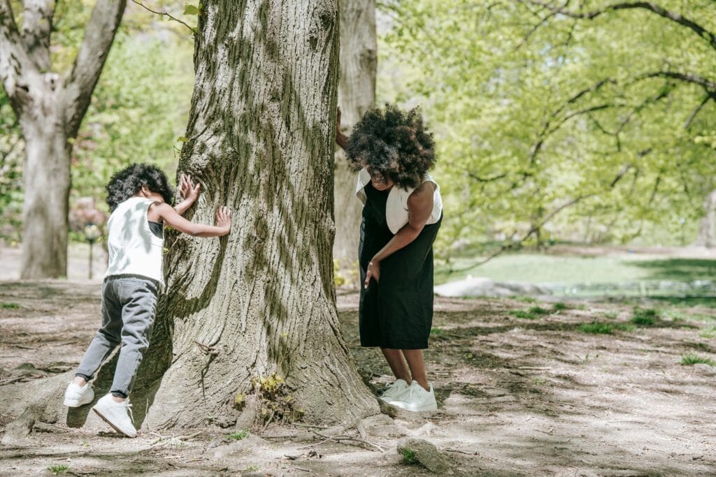 Hidden Object Puzzles. Two children playing hide-and-seek outdoors, with one leaning against a large tree trunk and the other peeking around it in a park filled with green foliage.
