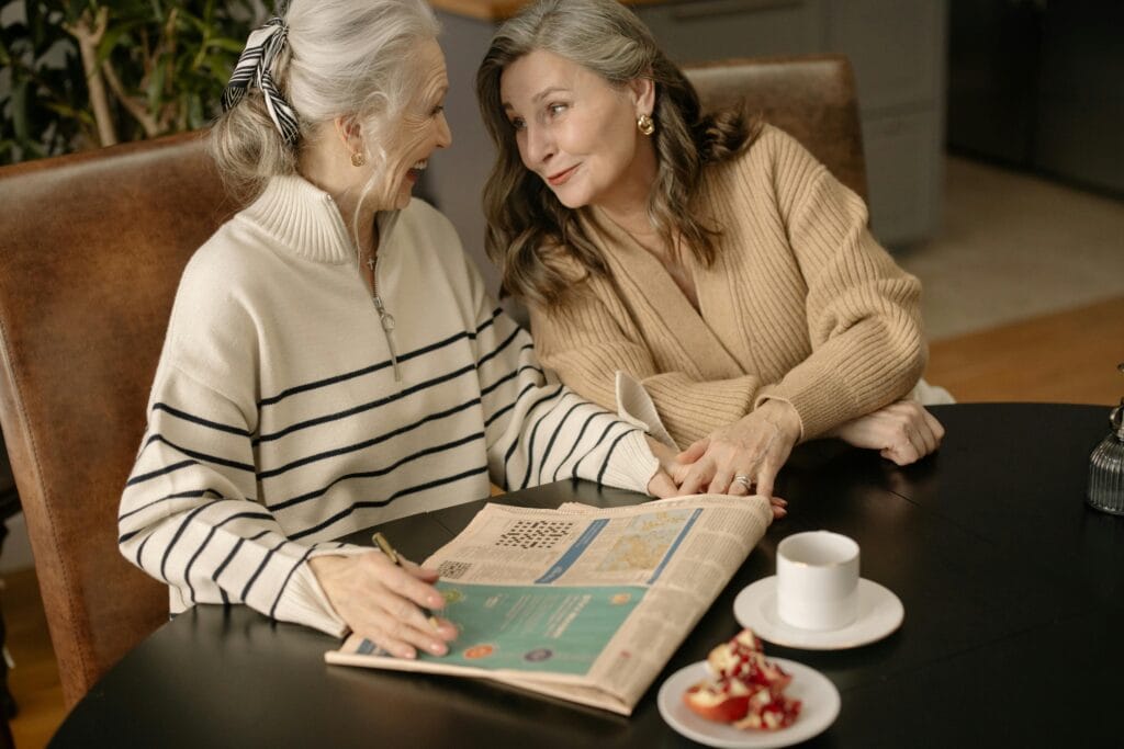  Spot the Difference. Two older women sitting at a table, smiling warmly at each other while holding hands, with a newspaper, cup of coffee, and a plate of pomegranate seeds in front of them.