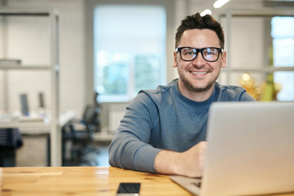 Featuring puzzle flipbook.  A smiling man wearing glasses and a blue sweatshirt sitting at a desk with a laptop in front of him inside a modern office setting.