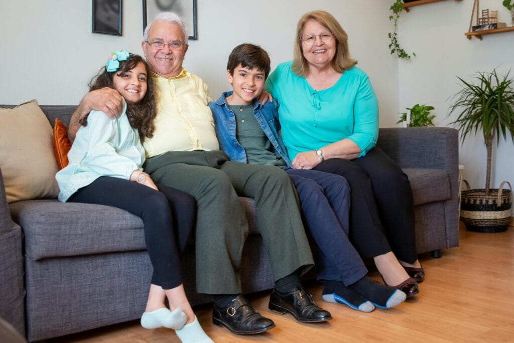 Featuring Spot the Difference for everyone. Smiling grandparents sitting on a sofa with their two young grandchildren, all seated closely together in a cozy living room with houseplants and wall shelves in the background.