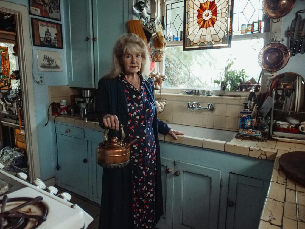 Spot the Difference: Elderly woman standing in a vintage kitchen, holding a copper kettle, with stained glass windows and retro decor in the background.