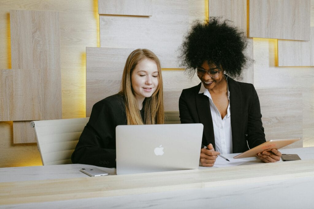 Spot the Difference Books. Two businesswomen collaborating at a desk, smiling as they review work on a laptop and tablet in a modern office setting.
