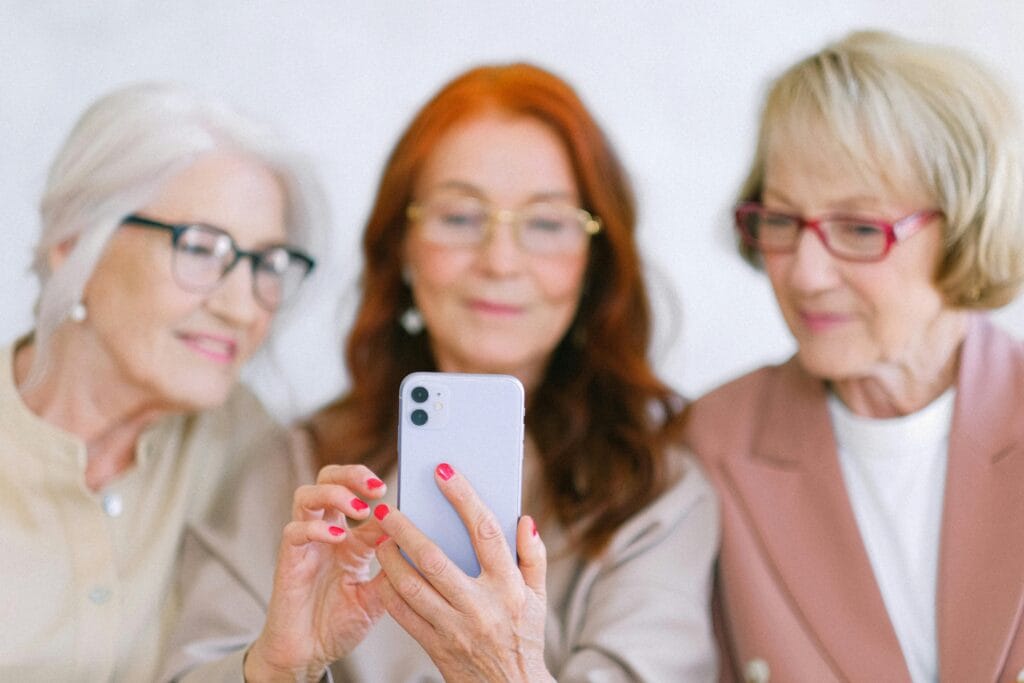Spot the Difference for Seniors. Three elderly women wearing glasses, smiling and gathered closely while looking at a smartphone, with the focus on the phone in the foreground