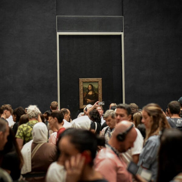 A large crowd gathers in front of the Mona Lisa painting, which is displayed on a black wall. The atmosphere is bustling, with people photographing it.