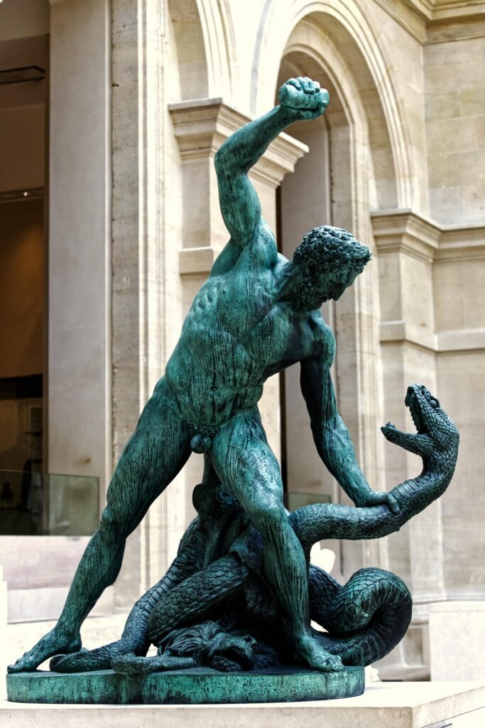 A large green bronze statue of a muscular man, likely Hercules, engaged in a dynamic struggle with a giant serpent within a museum courtyard.
