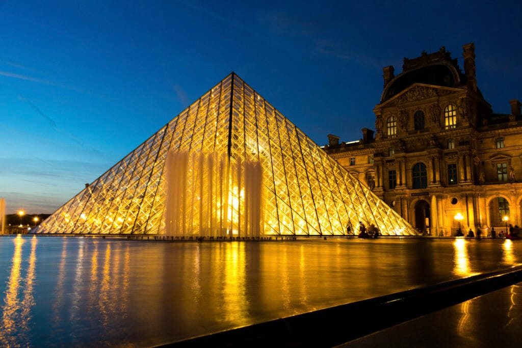 Illuminated glass pyramid at the Louvre Museum in Paris against a deep blue twilight sky, reflecting in a serene water pool, exuding elegance and grandeur.