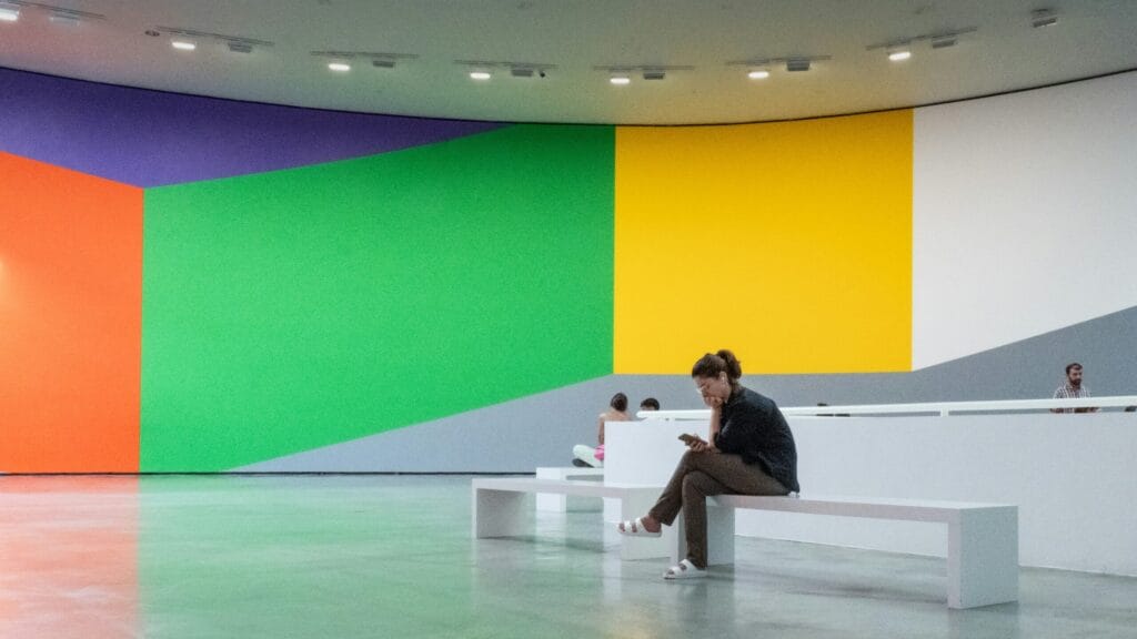 Museum Visit. A woman sits on a minimalist white bench in a modern art gallery, looking at her phone. In the background, a massive curved wall features a vibrant mural with bold geometric blocks of orange, purple, green, yellow, and white.