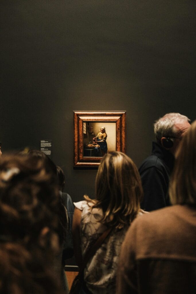 Visiting Museums. A crowd of museum visitors gathered around the famous, small-scale painting "The Milkmaid" by Johannes Vermeer on a dark gallery wall.