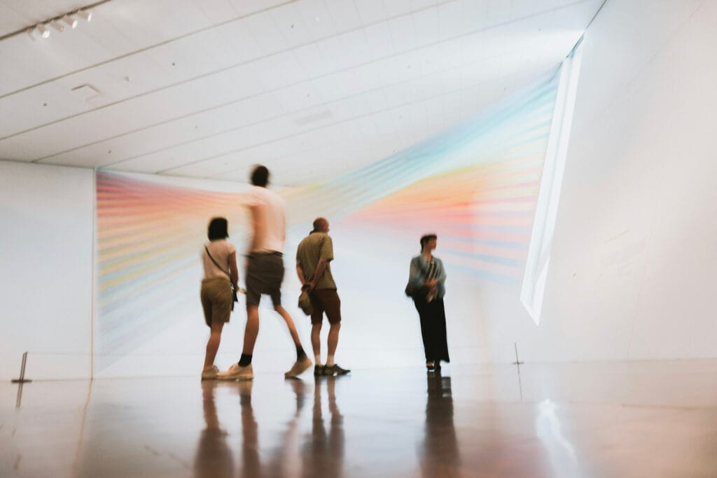 museum. A group of four people admire a colorful, rainbow light installation on a white gallery wall. The atmosphere feels calm and contemplative.