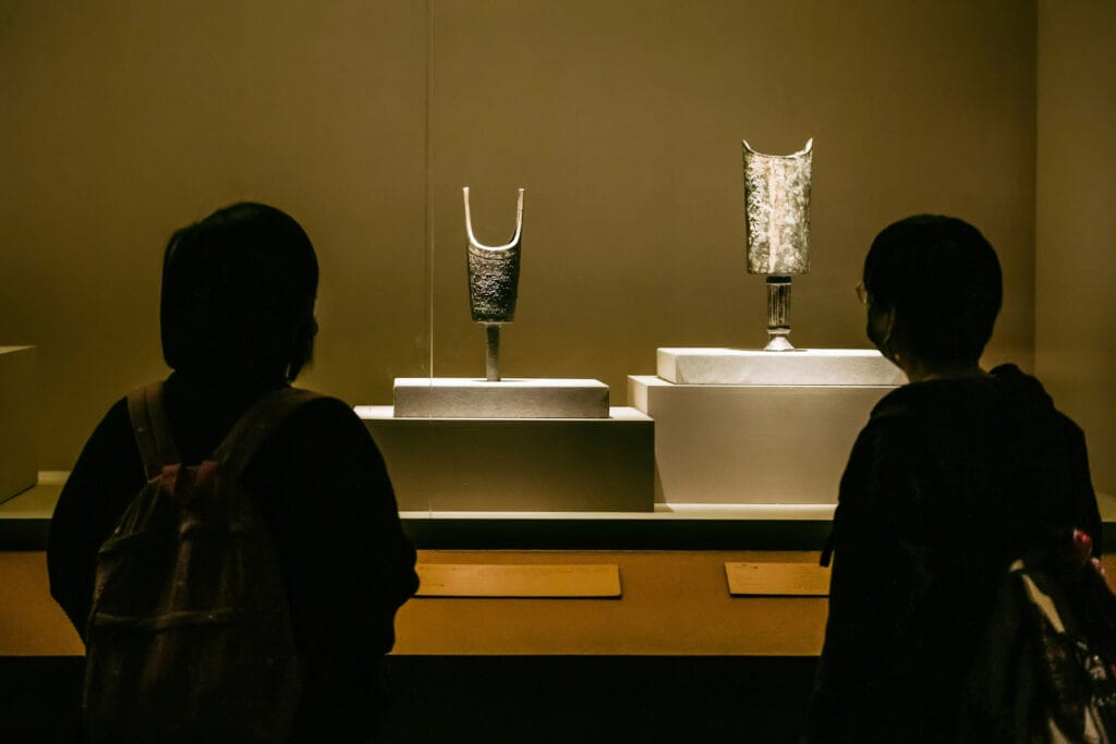 Silhouetted views of two people from behind as they observe ancient metallic artifacts displayed inside a glass case at a museum. The artifacts, which appear to be antique vessels or tools, are mounted on white pedestals under warm, dramatic lighting.