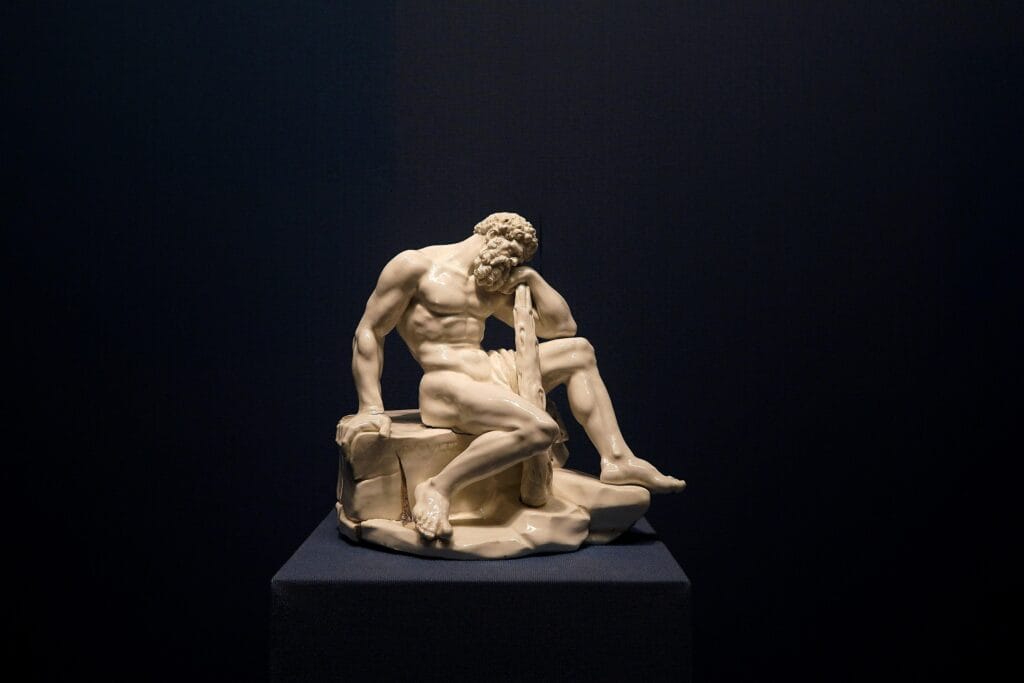 Visiting a Museum. A small, white classical sculpture of a muscular bearded man sitting and leaning on a club, displayed on a blue pedestal against a dark background.